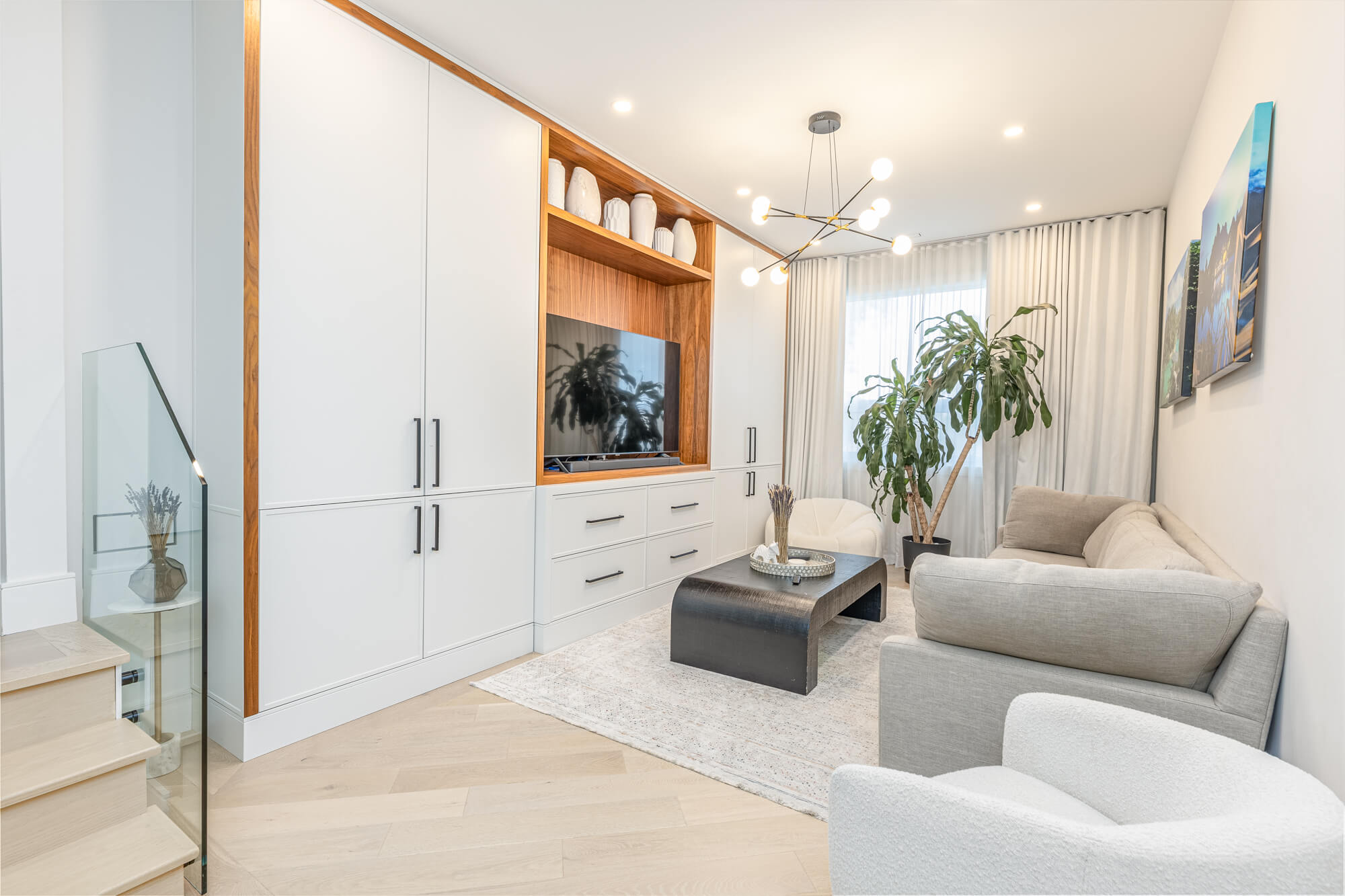 Living room in Ontario Airbnb rental — custom built-in TV wall unit with walnut accents, herringbone hardwood floors, sputnik chandelier and glass staircase railing