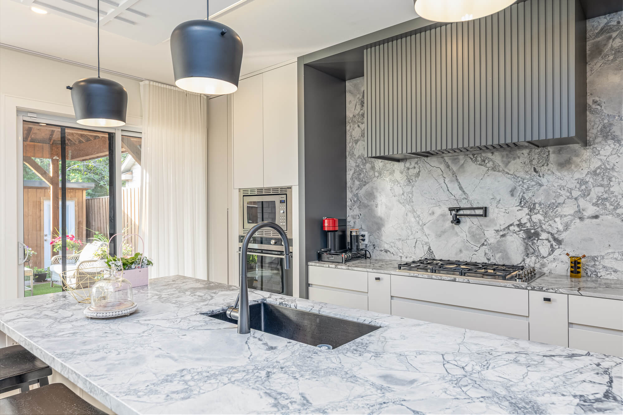 Modern Airbnb kitchen in Ontario — full-height marble backsplash, waterfall island with black undermount sink, slatted range hood and backyard access through sliding doors